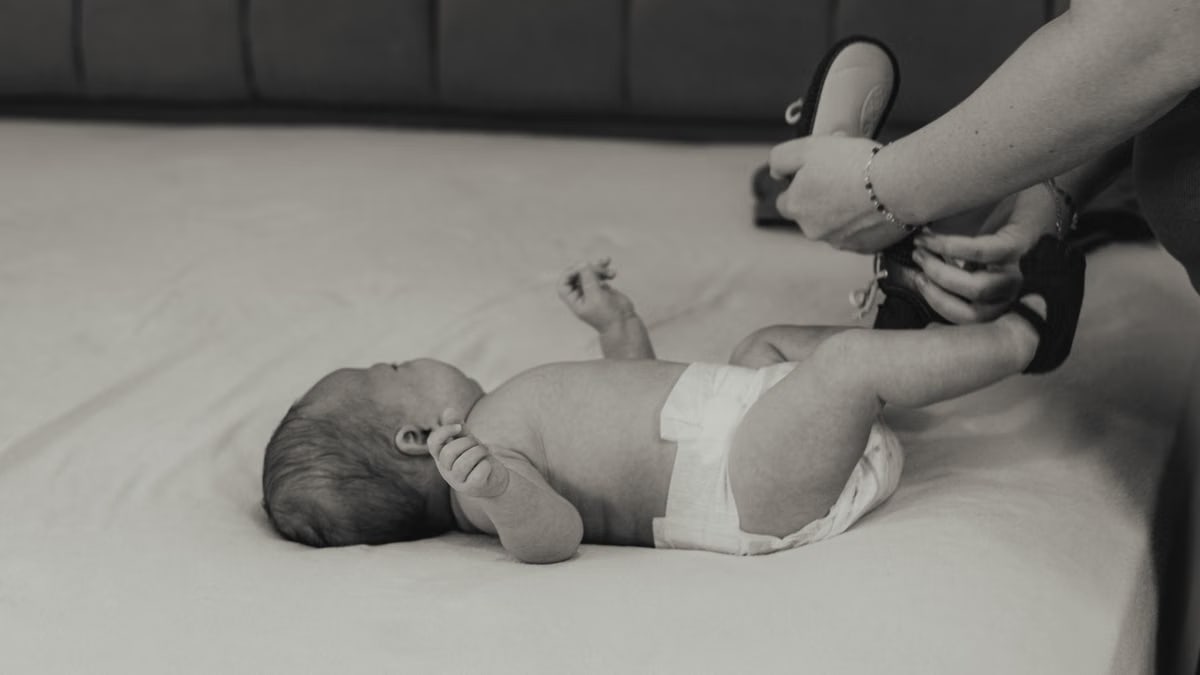 A mother lovingly dresses her newborn baby lying on a bed in an intimate black and white setting.